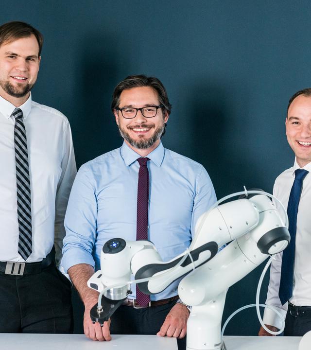 Drei Männer stehen hinter einem Roboterarm und lachen in die Kamera.