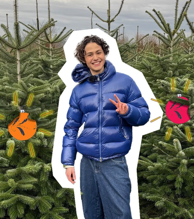 Reporter Gianni steht mit einer blauen Jacke gekleidet zwischen vielen Tannenbäumen auf einem Feld, neben ihn sieht man Christbaumkugeln.