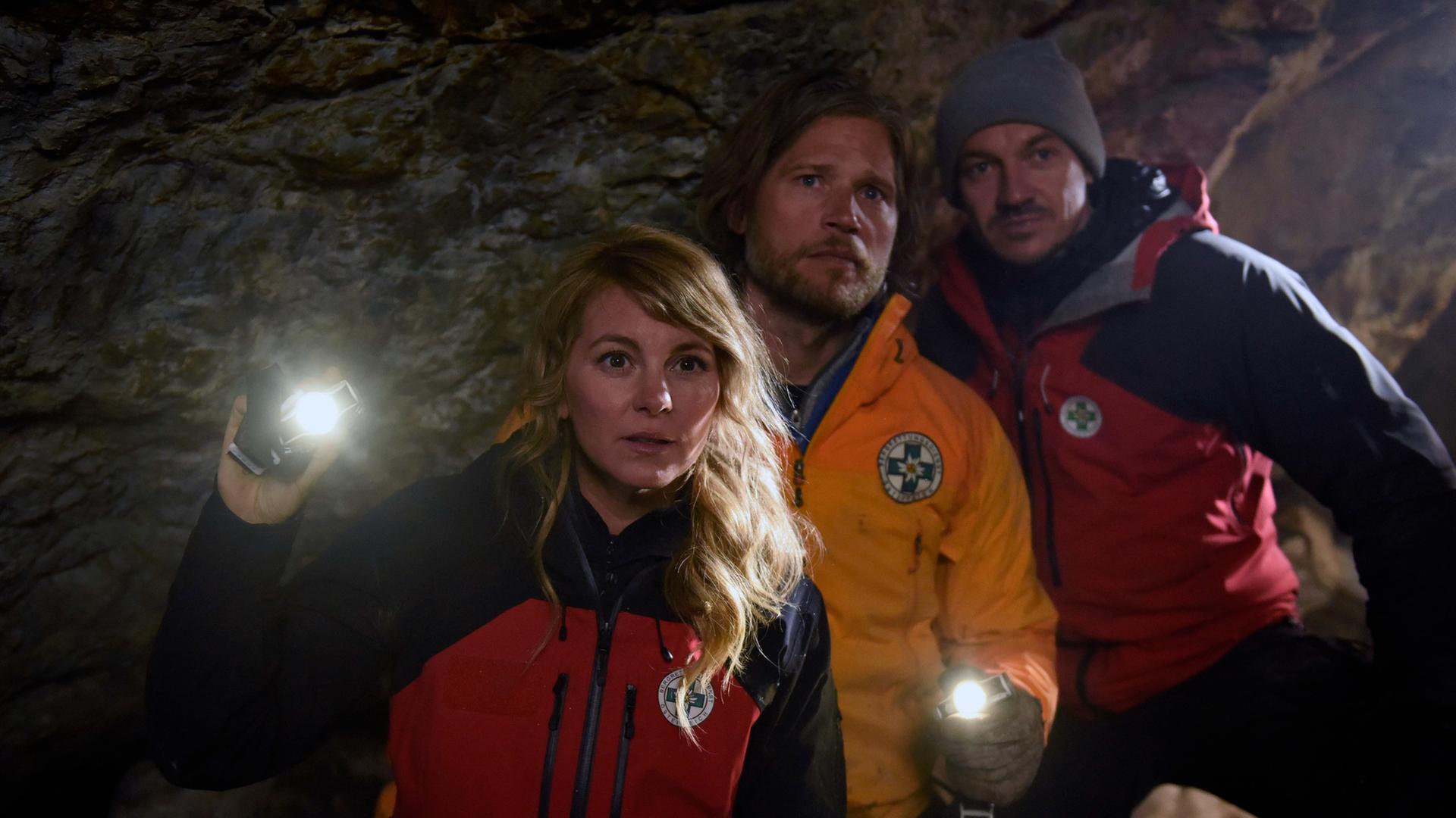 "Die Bergretter: Giftiges Erbe": In einer dunklen Höhle stehen die Bergretter Katharina (Luise Bähr), Markus (Sebastian Ströbel) und Tobi (Markus Brandl). Markus und Katharina leuchten mit Taschenlampen in die Höhle hinein. Alle blicken erwartungsvoll.