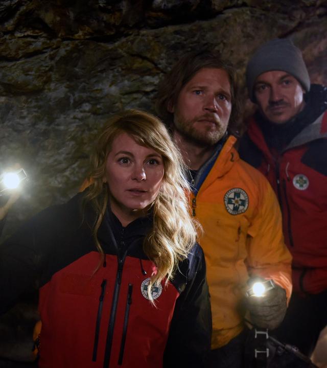 "Die Bergretter: Giftiges Erbe": In einer dunklen Höhle stehen die Bergretter Katharina (Luise Bähr), Markus (Sebastian Ströbel) und Tobi (Markus Brandl). Markus und Katharina leuchten mit Taschenlampen in die Höhle hinein. Alle blicken erwartungsvoll.