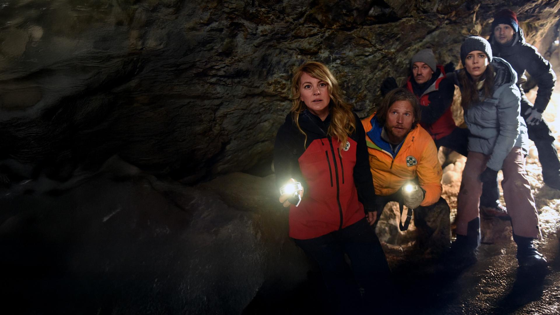 "Die Bergretter - Giftiges Erbe (2)": In einem dunklen Höhleneingang stehen die Bergretter Katharina (Luise Bähr), Markus (Sebastian Ströbel) und Tobi (Markus Brandl) sowie Laura Brandtner (Josepha Walter) und Max (Doğa Gürer). Markus und Katharina leuchten mit Taschenlampen in die Höhle hinein. Alle blicken erwartungsvoll.