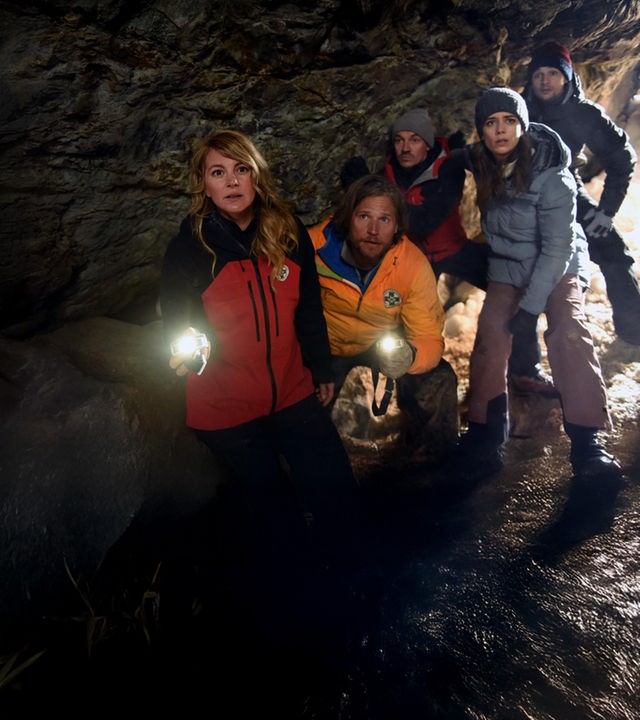 "Die Bergretter - Giftiges Erbe (2)": In einem dunklen Höhleneingang stehen die Bergretter Katharina (Luise Bähr), Markus (Sebastian Ströbel) und Tobi (Markus Brandl) sowie Laura Brandtner (Josepha Walter) und Max (Doğa Gürer). Markus und Katharina leuchten mit Taschenlampen in die Höhle hinein. Alle blicken erwartungsvoll.