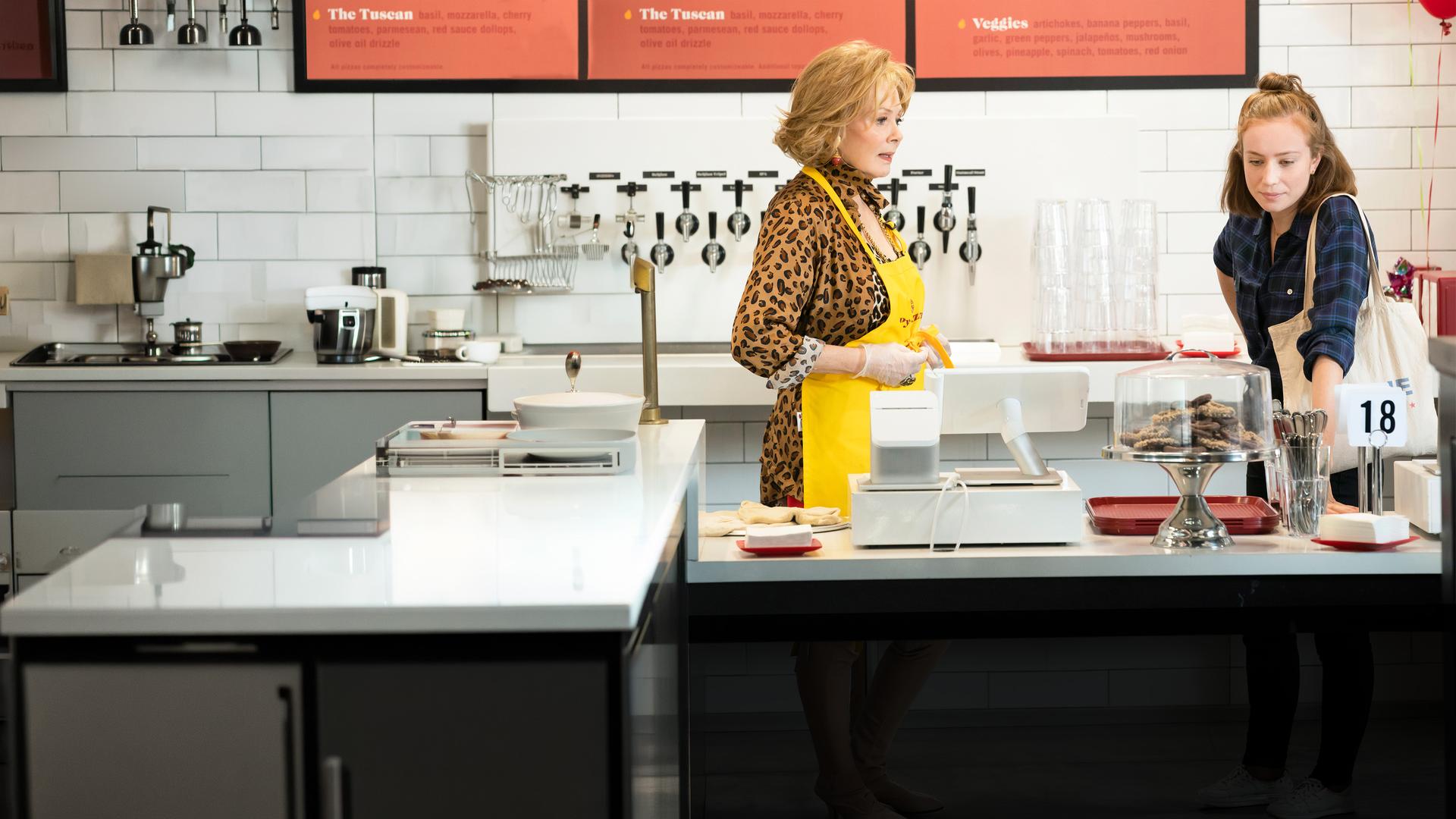 "Hacks – Gig ist eben Gig": Deborah (Jean Smart) trägt eine gelbe Schürze und steht hinter einem Tresen. Mit ernstem Blick wendet sie sich Ava (Hannah Einbinder) zu, die neben ihr steht und nachdenklich auf die Tischplatte des Tresens schaut. Im Hintergrund sind weiße Fliesen, Zapfhähne und Menütafeln zu sehen.