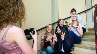 Auf einer hellen Treppe im ZDF-Landesstudio Schwerin haben sich junge Teilnehmerinnen des Girls’ Day 2026 versammelt, sitzen dicht beieinander, lachen in die Kamera und werden gerade von einer weiteren Teilnehmerin fotografiert