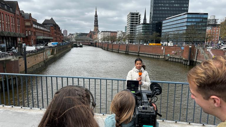 Mehrere Jugendliche stehen auf einer Brücke über einen Kanal in Hamburg und filmen mit einer professionellen Kamera ein Mädchen, das als Reporterin vor der Linse spricht. Im Hintergrund sind Backsteingebäude, moderne Bürohäuser und eine der Hamburger Michel (Kirche) unter bewölktem Himmel zu sehen