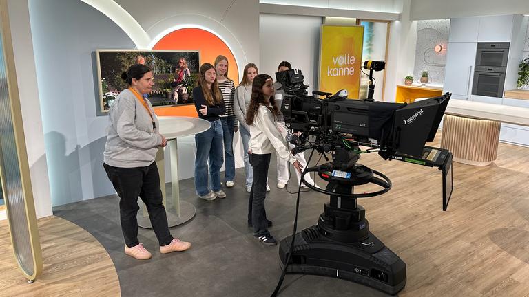 Mehrere jugendliche Mädchen stehen im Studio der ZDF-Sendung "Volle Kanne" und beobachten, wie eine Teilnehmerin eine professionelle Fernsehkamera bedient. Die Gruppe erhält im Rahmen des Girls’ Day 2026 praktische Einblicke in die Studioarbeit und Kameratechnik