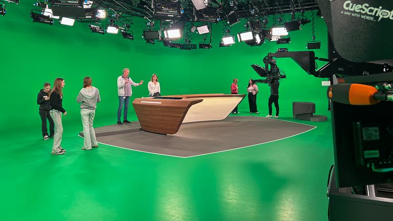 Eine Gruppe von Mädchen steht im ZDF-Nachrichtenstudio vor einer großen Greenscreen-Fläche und rund um einen Nachrichtentisch. Studioleuchten und Kameratechnik sind sichtbar, während sie beim Girls’ Day 2026 Einblicke in die Nachrichtenproduktion erhalten.