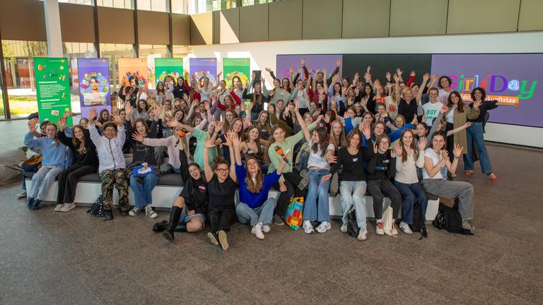 Zahlreiche Mädchen und junge Frauen versammeln sich für ein Gruppenfoto im ZDF und lachen sowie winken in die Kamera. Hinter ihnen hängen farbenfrohe Aktionsplakate zum Girls’ Day 2026, die die gemeinsame Veranstaltung sichtbar machen