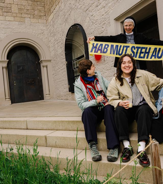 Drei junge Frauen sitzen auf einer Treppe mit Getränk in der Hand, hinter ihnen seht eine Ordensschwester mit dem Katholikentags -Schal mit dem Motto "Hab Mut, steh auf!"