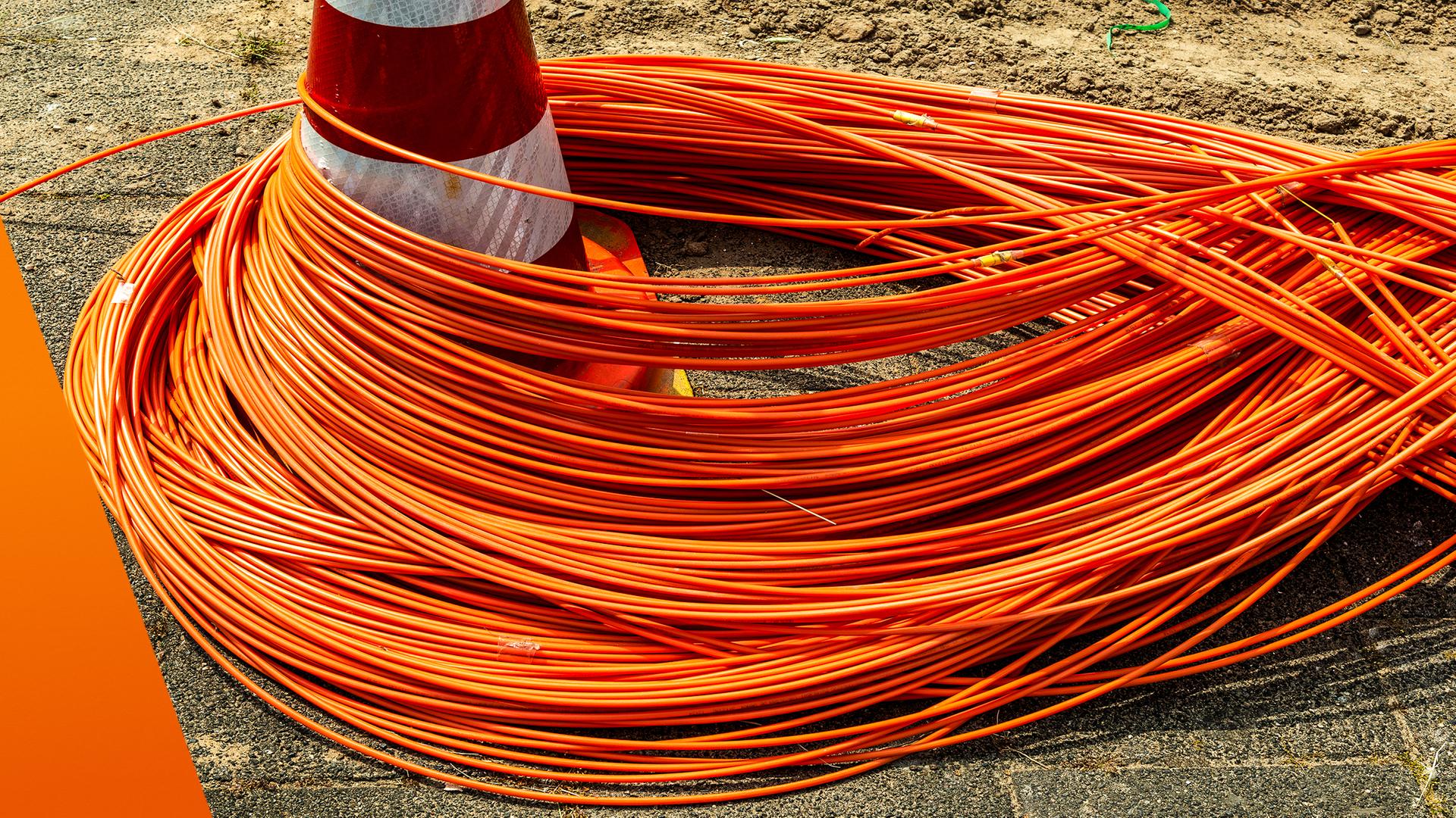 Auf einer Baustelle liegen zahlreiche orangefarbene Glasfaserkabel in dichten Schlaufen auf dem Boden. Ein rot-weißer Verkehrskegel steht in der Mitte und wird teilweise von den Kabeln umschlungen. Der Untergrund besteht aus Asphalt und Erde, was auf laufende Bauarbeiten für den Netzausbau hinweist.