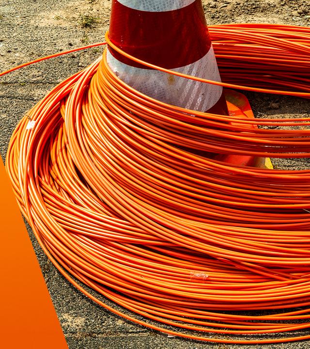 Auf einer Baustelle liegen zahlreiche orangefarbene Glasfaserkabel in dichten Schlaufen auf dem Boden. Ein rot-weißer Verkehrskegel steht in der Mitte und wird teilweise von den Kabeln umschlungen. Der Untergrund besteht aus Asphalt und Erde, was auf laufende Bauarbeiten für den Netzausbau hinweist.