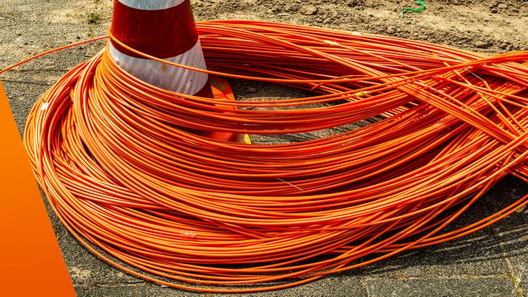 Auf einer Baustelle liegen zahlreiche orangefarbene Glasfaserkabel in dichten Schlaufen auf dem Boden. Ein rot-weißer Verkehrskegel steht in der Mitte und wird teilweise von den Kabeln umschlungen. Der Untergrund besteht aus Asphalt und Erde, was auf laufende Bauarbeiten für den Netzausbau hinweist.