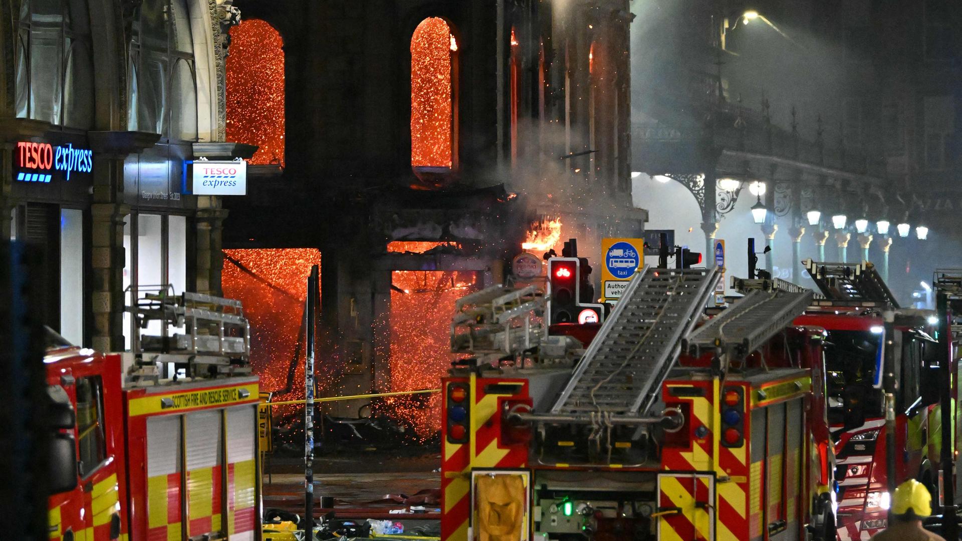 Gebäude steht nachts unter Flammen, davor parken Feuerwehrautos.