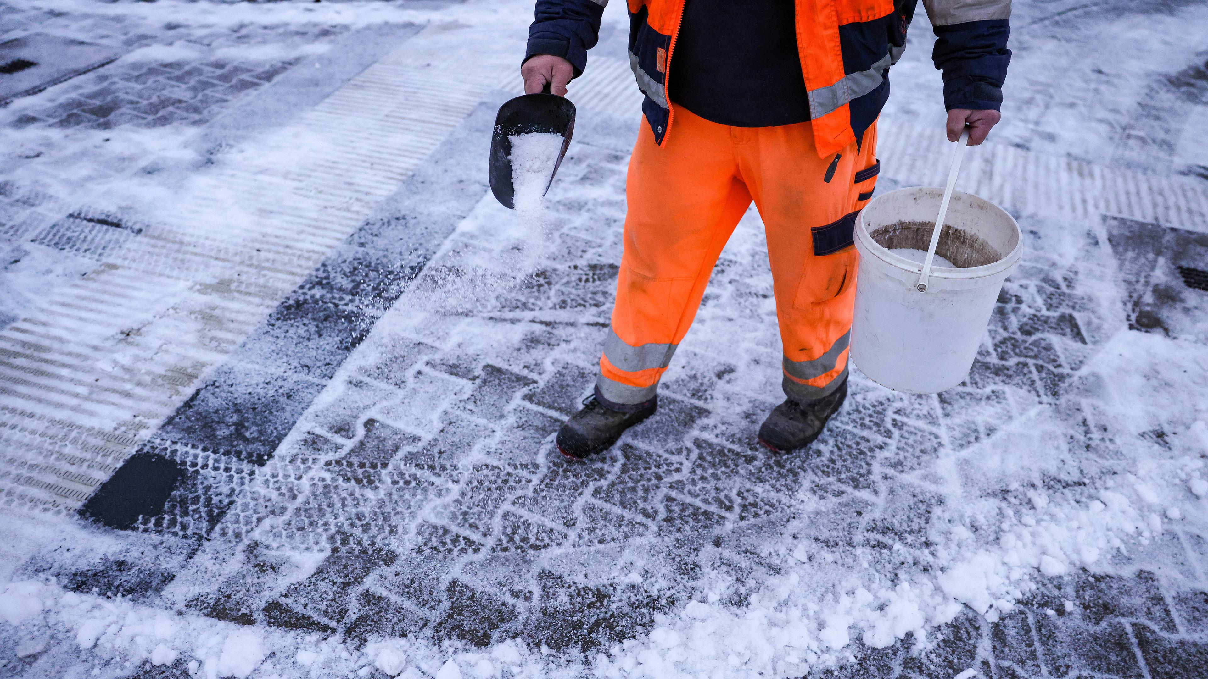 Ein Mitarbeiter des Winterdienstes verteilt Streusalz bei Regen und Schneeglätte auf den Fußweg an einer Straßenkreuzung.