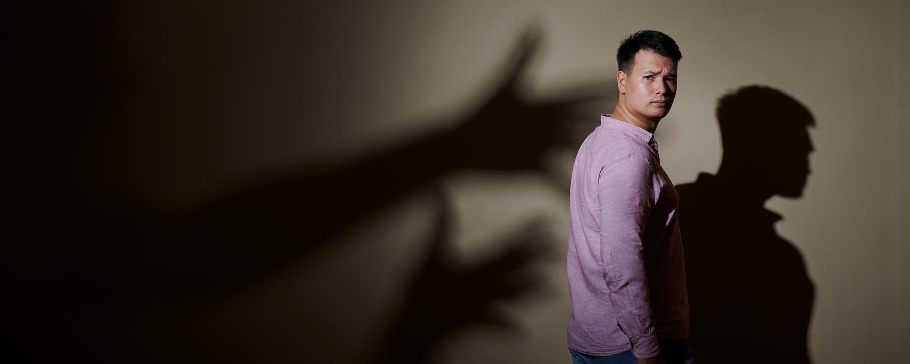 Max Leon steht in Jeans und rosa Hemd seitlich vor einer Wand. Hinter ihm ist sein Schatten an der Wand und Schattenhände, die nach ihm greifen wollen.