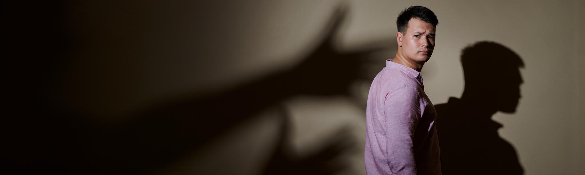 Max Leon steht in Jeans und rosa Hemd seitlich vor einer Wand. Hinter ihm ist sein Schatten an der Wand und Schattenhände, die nach ihm greifen wollen.