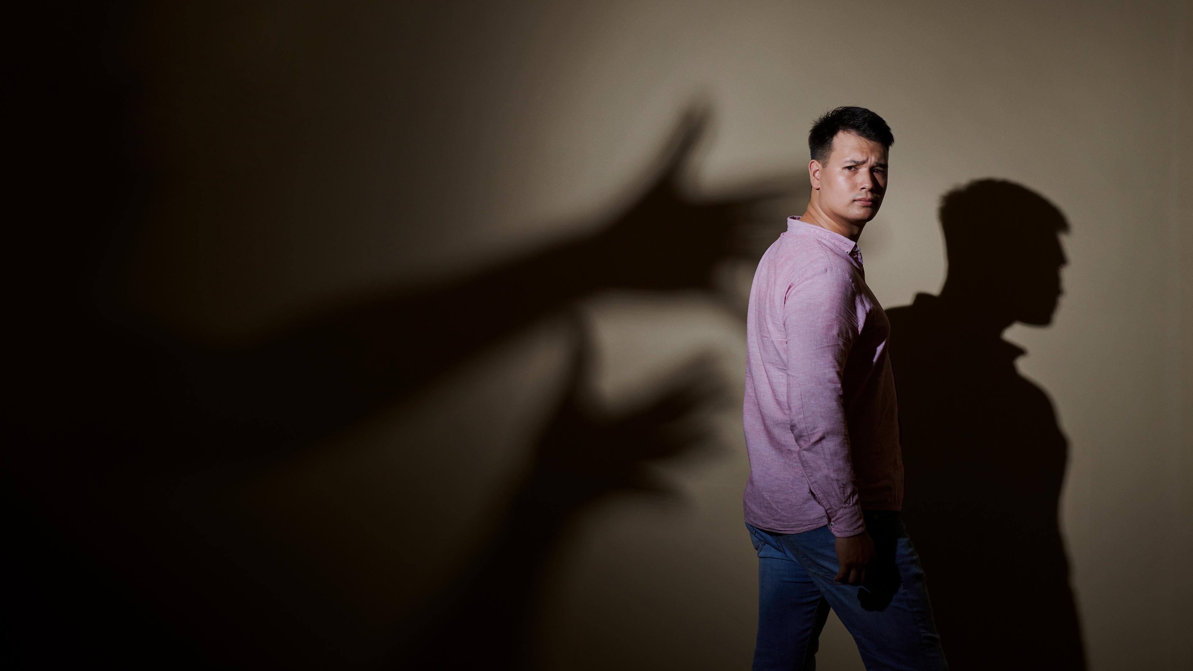 Max Leon steht in Jeans und rosa Hemd seitlich vor einer Wand. Hinter ihm ist sein Schatten an der Wand und Schattenhände, die nach ihm greifen wollen.