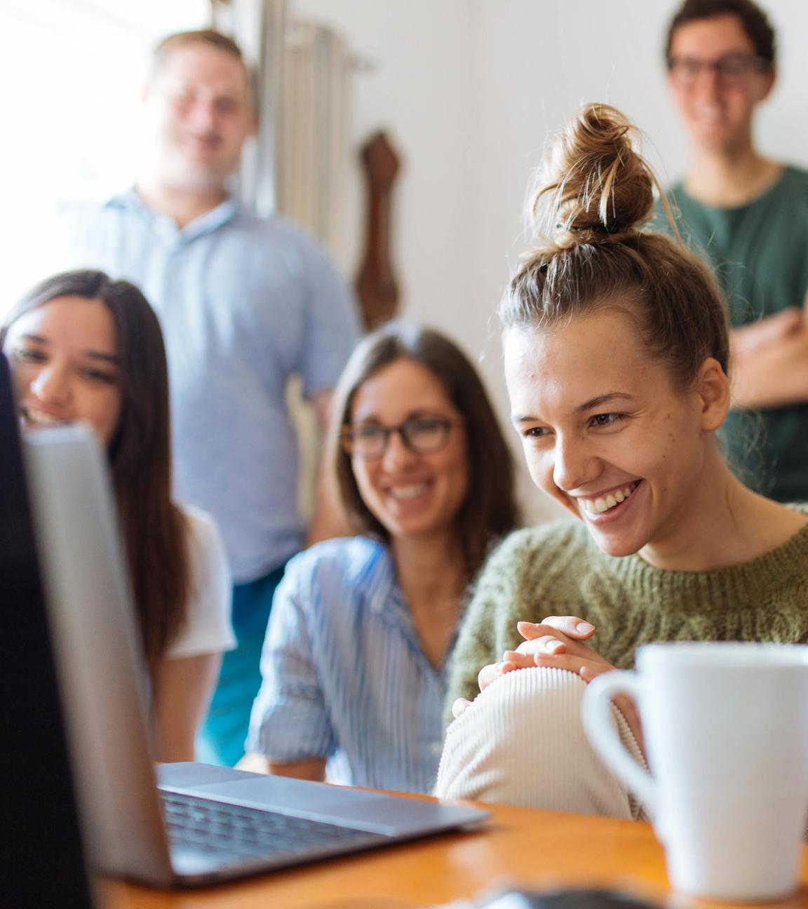 Vier junge Frauen lachen vor einem Laptop sitzend, im Hintergrund unscharf zwei junge Männer stehend