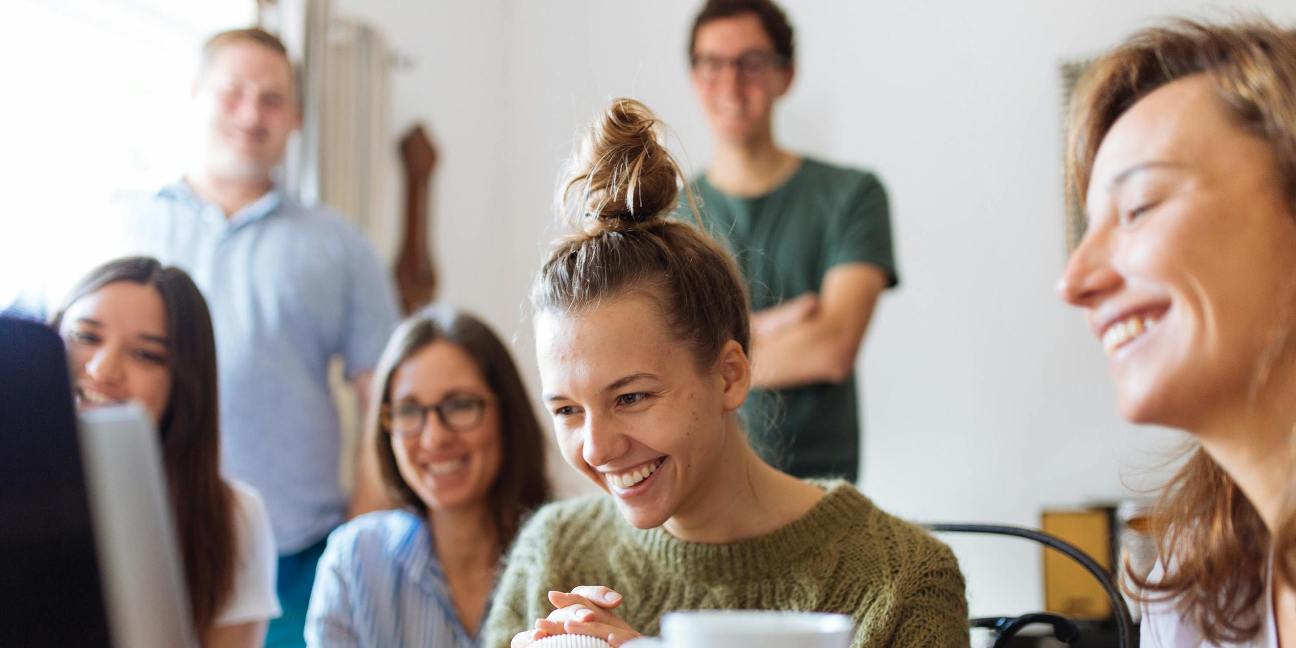 Vier junge Frauen lachen vor einem Laptop sitzend, im Hintergrund unscharf zwei junge Männer stehend