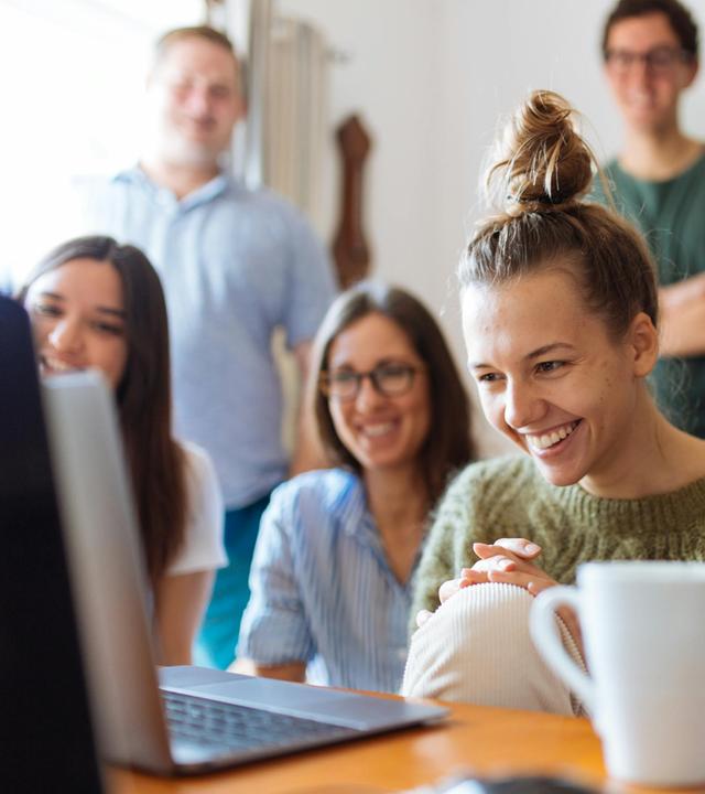 Vier junge Frauen lachen vor einem Laptop sitzend, im Hintergrund unscharf zwei junge Männer stehend