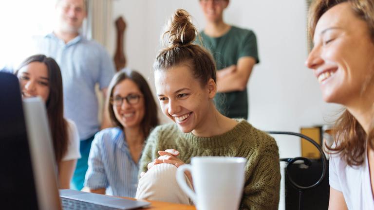 Vier junge Frauen lachen vor einem Laptop sitzend, im Hintergrund unscharf zwei junge Männer stehend