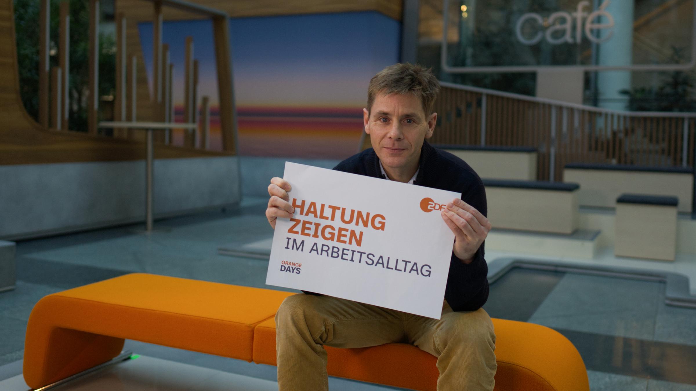 ZDF-Mitarbeiter des ZDF-Studios in Berlin hält Schild zum Aktionstag "Orange Days" mit der Aufschrift "Haltung zeigen im Arbeitsalltag" in die Kamera.