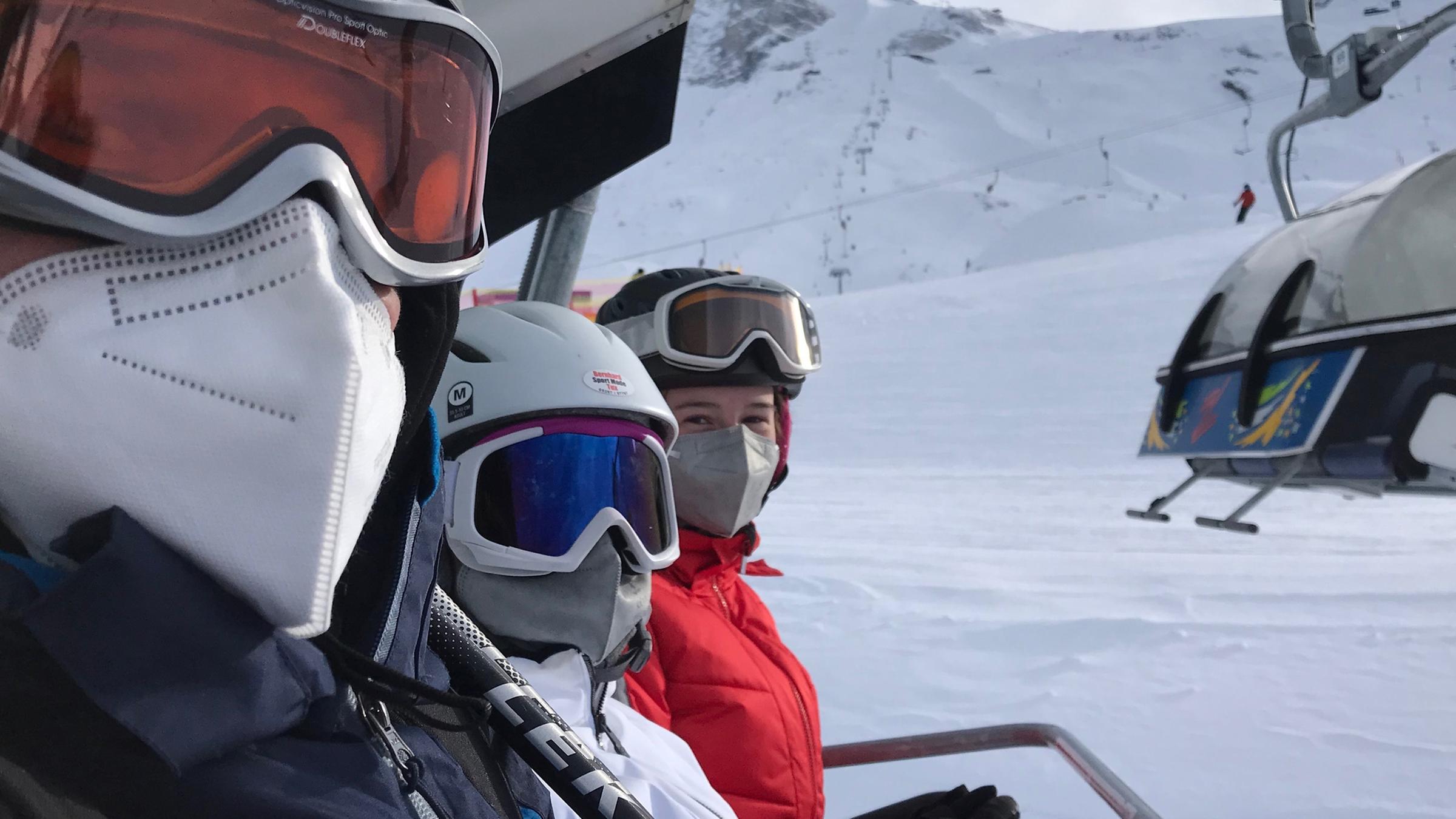 Drei Personen sitzen mit Skibrillen und FFP2-Maske nebeneinander im Sessellift und schauen in die Kamera.