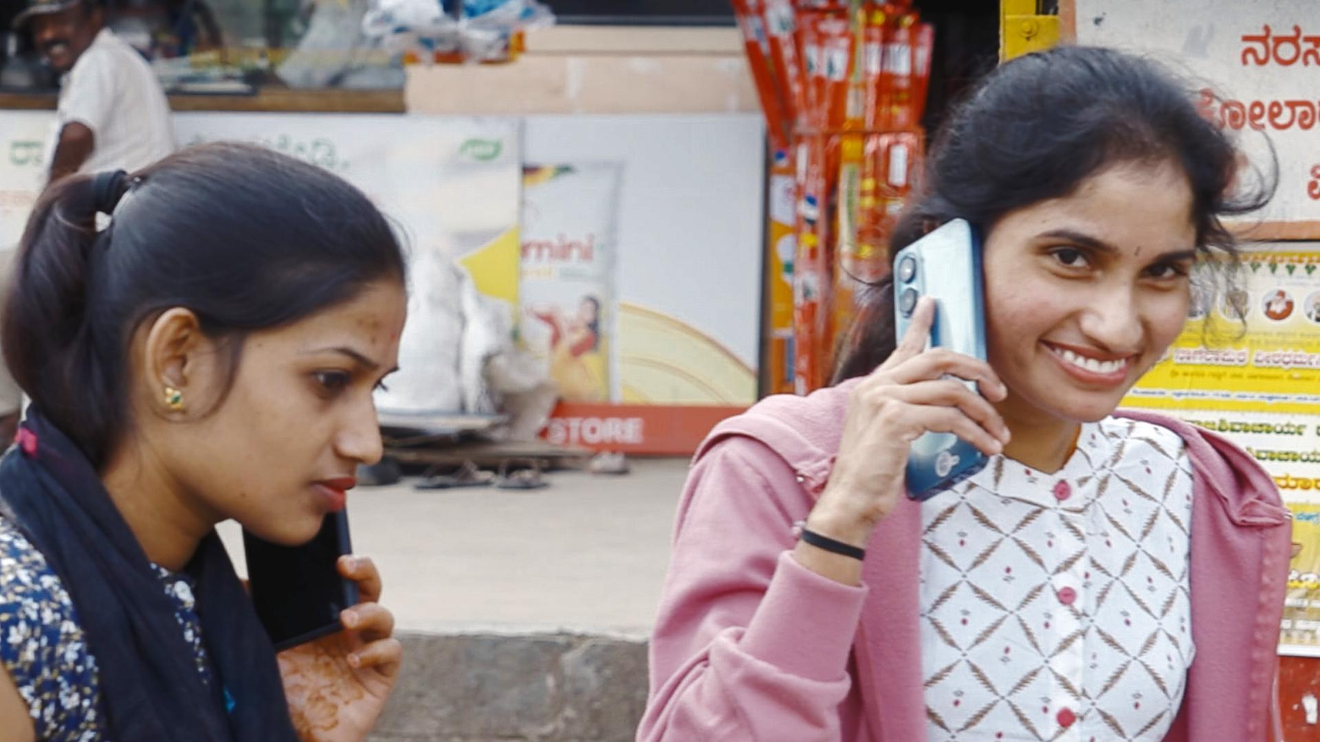 Zwei junge Frauen telefonieren mit ihren Smartphones auf der Straße in der Provinz Bangalore, eine lächelt in die Kamera.