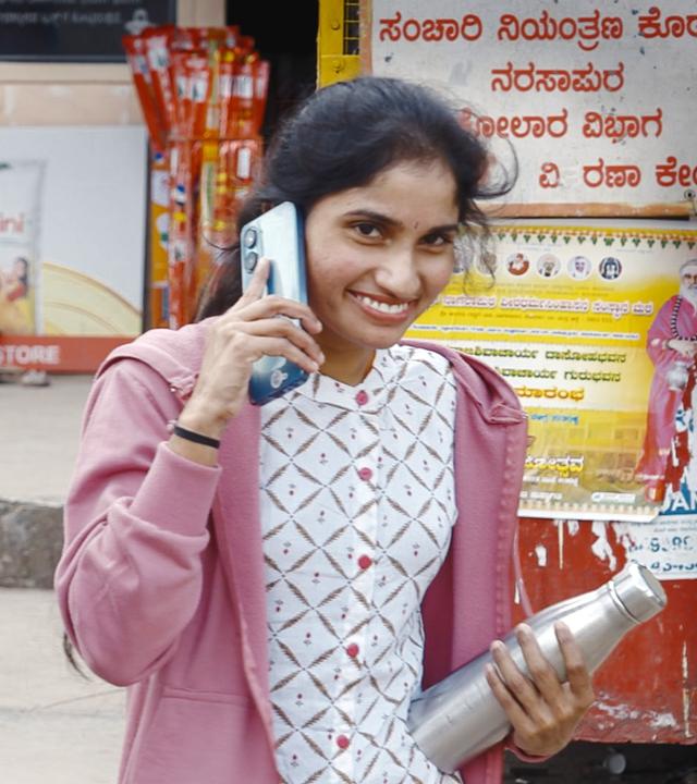 Zwei junge Frauen telefonieren mit ihren Smartphones auf der Straße in der Provinz Bangalore, eine lächelt in die Kamera.