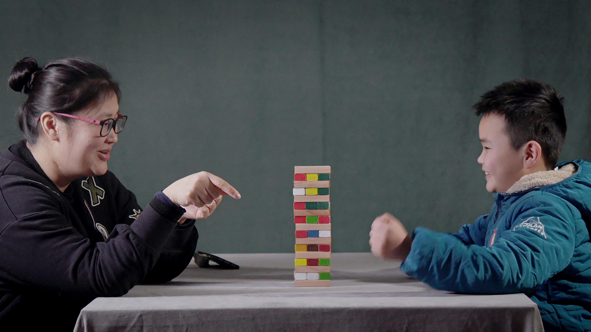 Auf einem Tisch steht ein Spiel aus Holzklötzchen. Die Mutter Qi Jingjing und ihr Sohn Liu Yufeng sitzen sich an dem Tisch gegenüber und spielen das Spiel Jenga mit den Holzklötzen. Beide haben Spaß dabei.