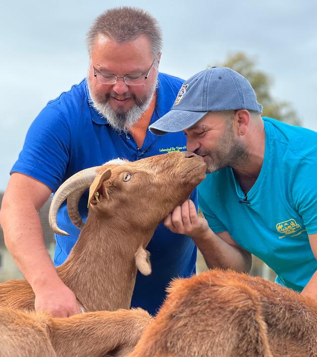 Peter und Daniel Rotter tragen die blauen T-Shirts mit dem Logo des Lebenshofs. Daniel küsst eine braune Ziege und Peter streichelt sie.
