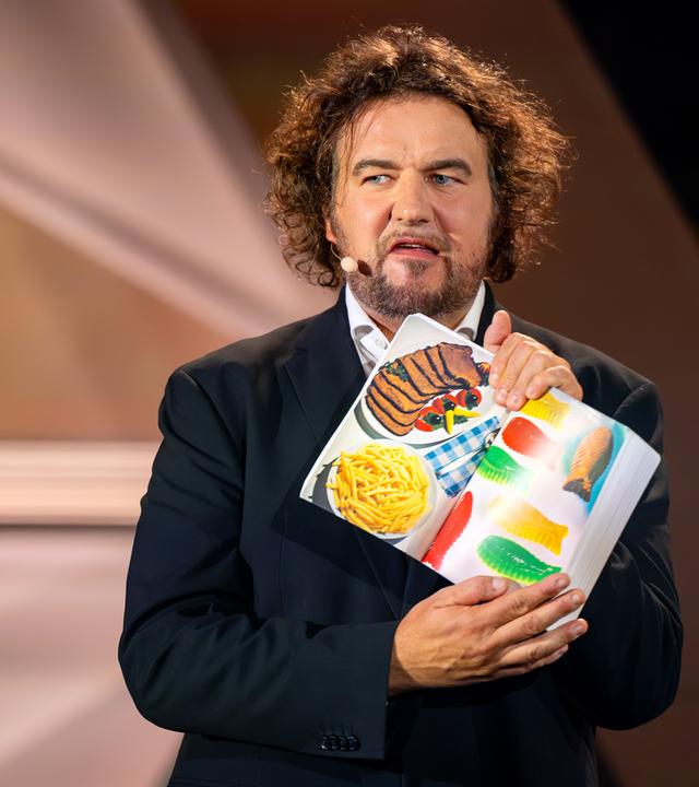 Götz Frittrang steht in der Mitte des Bildes. Er hält ein geöffnetes Buch in seiner Hand. Götz Frittrang trägt einen dunklen Anzug. Er verzieht sein Gesicht.