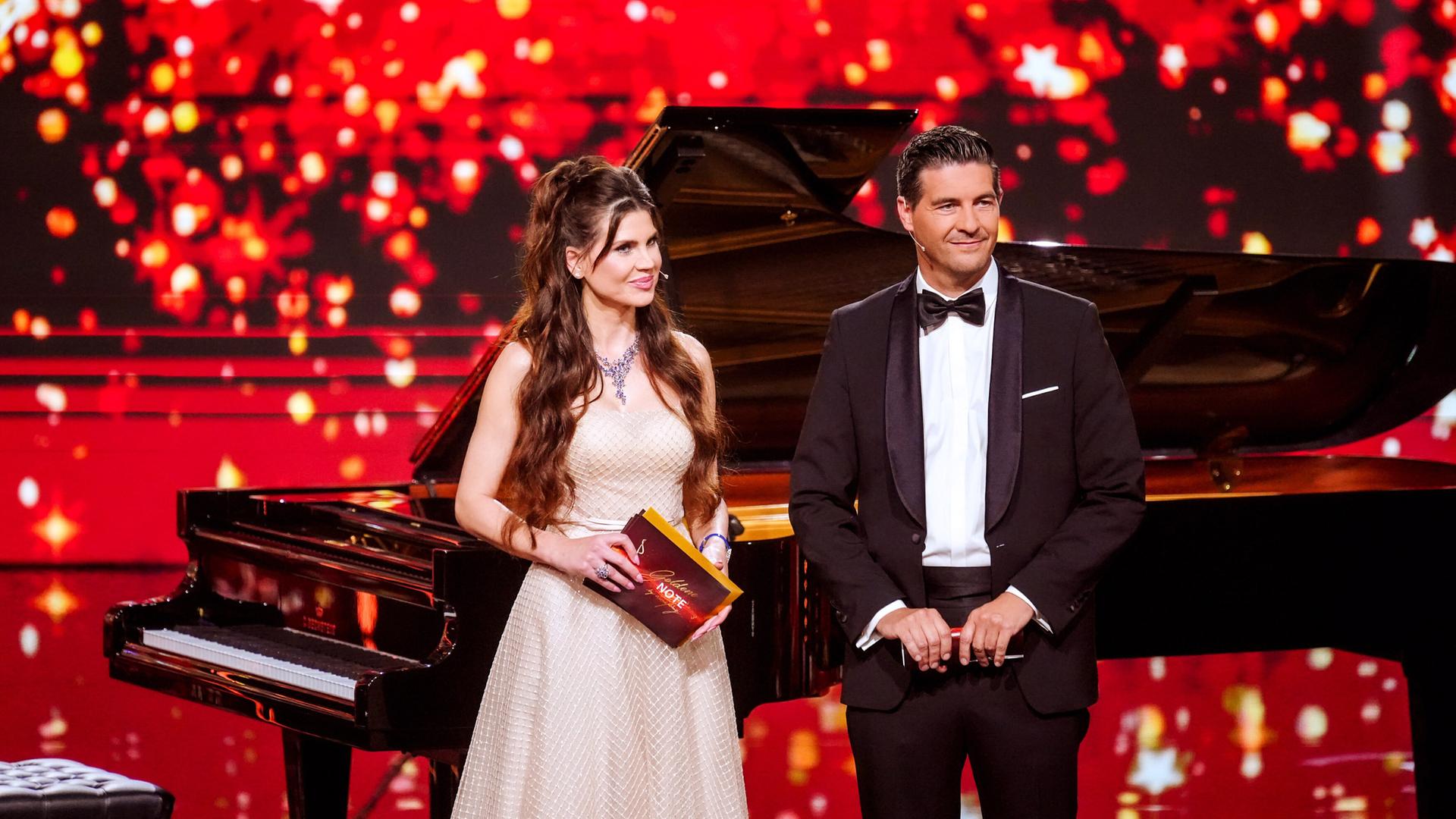Eine Frau und ein Mann in Abendrobe stehen auf einer rot beleuchteten Bühne mit einem Flügel im Hintergrund