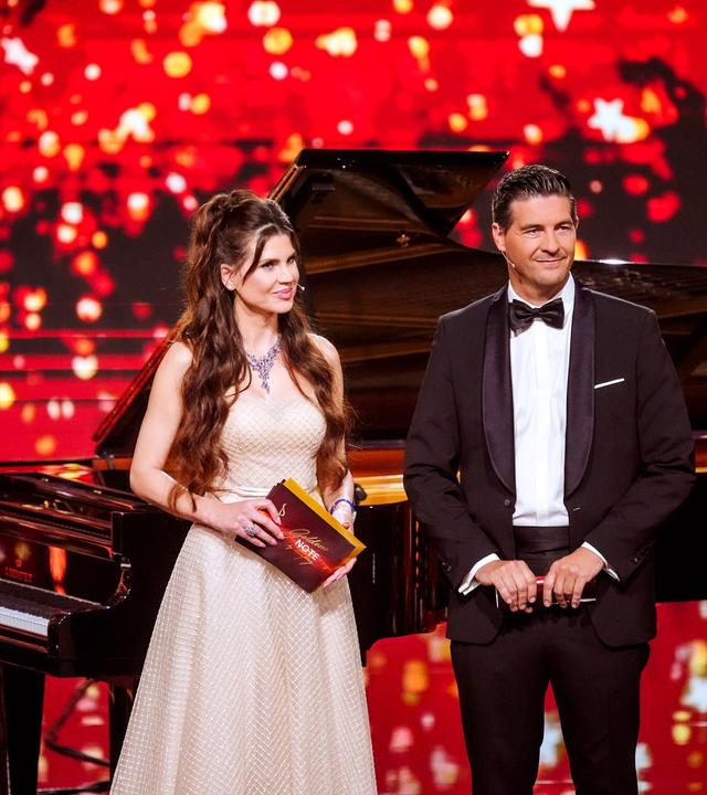 Eine Frau und ein Mann in Abendrobe stehen auf einer rot beleuchteten Bühne mit einem Flügel im Hintergrund