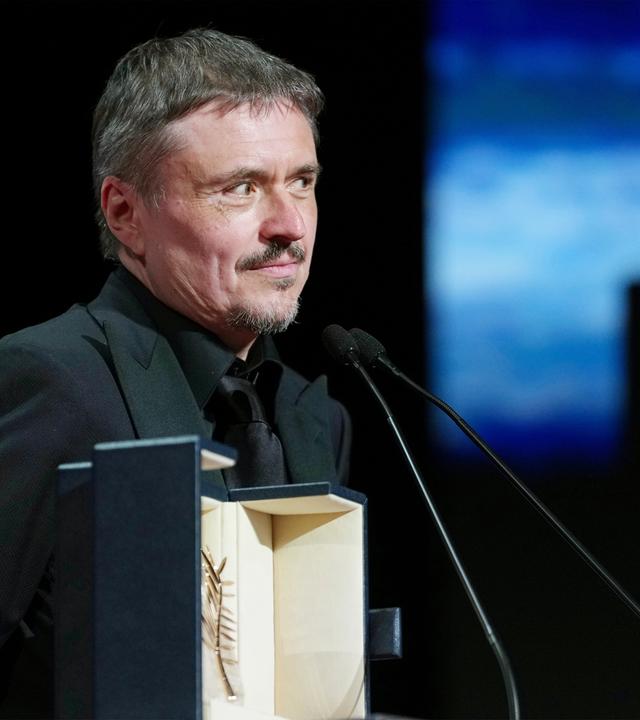 Ein Mann mit graumeliertem Bärtchen steht stolz hinter einem Rednerpult mit einem Kästchen, in dem eine goldene Palme präsentiert wird.
