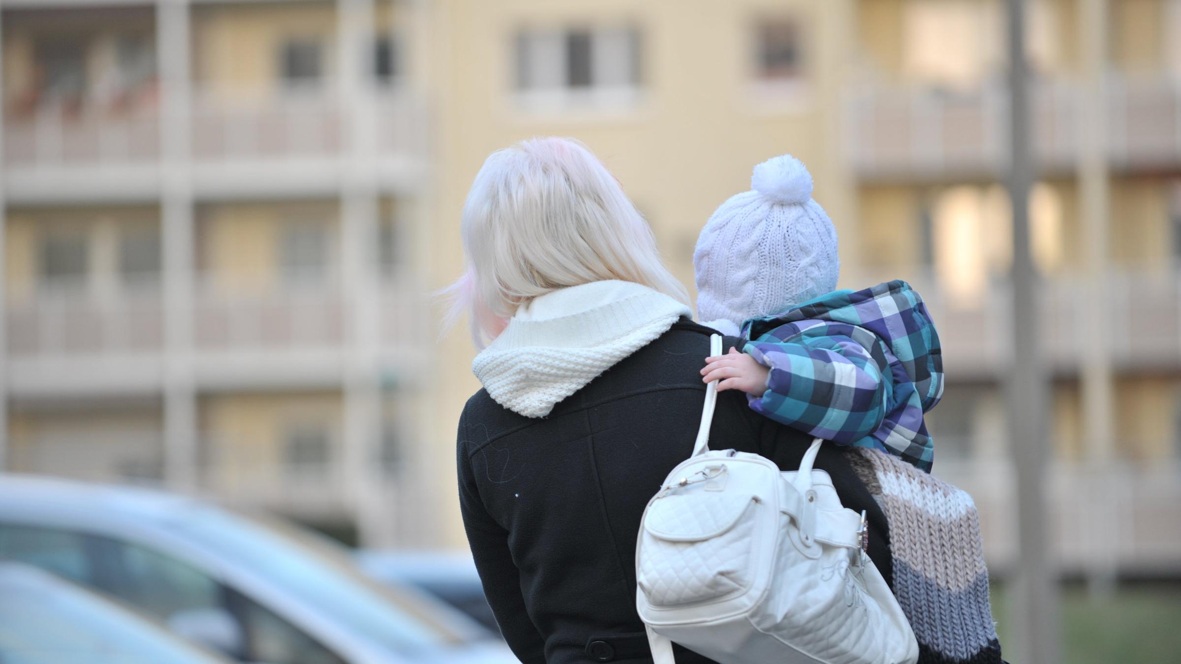 "Goldgrube Wohnungsnot – Spekulanten gegen Mieter": Mutter hält ihr kleines Kind auf dem Arm und schaut auf ein Wohnhaus.
