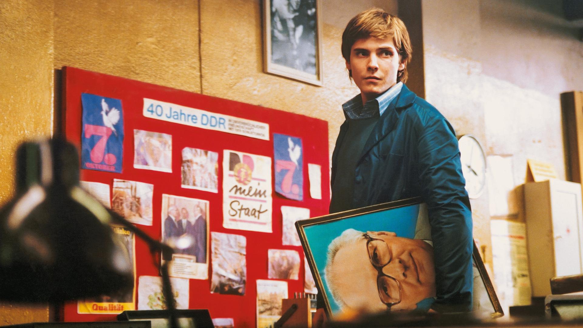 Alex blickt in einer Art Lagerhalle suchend um sich. Unter dem linken Arm trägt er ein Porträt Erich Honeckers. Im Hintergrund sieht man eine Fototafel mit der Überschrift „40 Jahre DDR“.