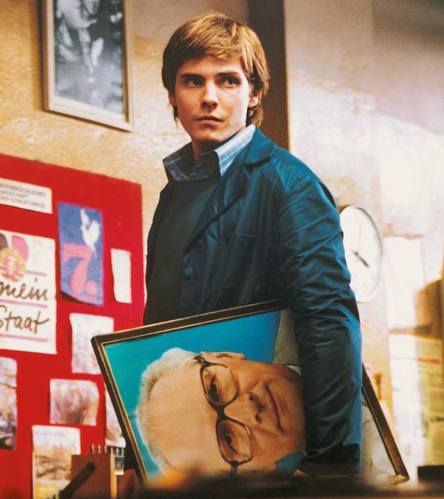 Alex blickt in einer Art Lagerhalle suchend um sich. Unter dem linken Arm trägt er ein Porträt Erich Honeckers. Im Hintergrund sieht man eine Fototafel mit der Überschrift „40 Jahre DDR“.