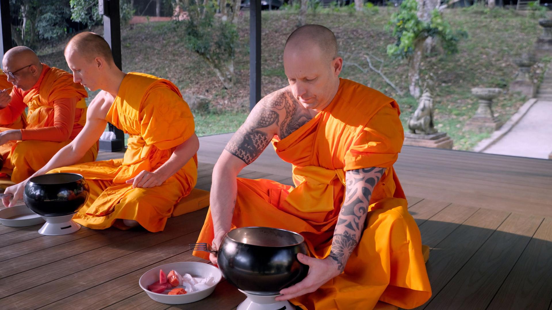 Das Bild zeigt einen buddhistischen Klosterraum im Freien, umgeben von Grünpflanzen. Im Vordergrund sitzen zwei Mönche in leuchtend orangefarbenen Roben auf Holzböden. Der Mönch auf der rechten Seite hat einen kalten Kopf und Tätowierungen auf dem linken Arm und ist dabei, einen schwarzen Almosenbehälter, der auf einer kleinen weißen Säule steht, in der Hand zu halten. Vor ihm liegt eine Schüssel mit geschnittenem Obst. Der Mönch auf der linken Seite trägt eine dunkelorange Robe und ist mit einer näheren Einstellung nicht vollständig sichtbar. Im Hintergrund sind Bäume und ein Fußweg zu erkennen, der leicht sichtbar ist. Die Stimmung wirkt friedlich und meditativer Natur.