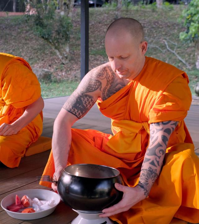 Das Bild zeigt einen buddhistischen Klosterraum im Freien, umgeben von Grünpflanzen. Im Vordergrund sitzen zwei Mönche in leuchtend orangefarbenen Roben auf Holzböden. Der Mönch auf der rechten Seite hat einen kalten Kopf und Tätowierungen auf dem linken Arm und ist dabei, einen schwarzen Almosenbehälter, der auf einer kleinen weißen Säule steht, in der Hand zu halten. Vor ihm liegt eine Schüssel mit geschnittenem Obst. Der Mönch auf der linken Seite trägt eine dunkelorange Robe und ist mit einer näheren Einstellung nicht vollständig sichtbar. Im Hintergrund sind Bäume und ein Fußweg zu erkennen, der leicht sichtbar ist. Die Stimmung wirkt friedlich und meditativer Natur.
