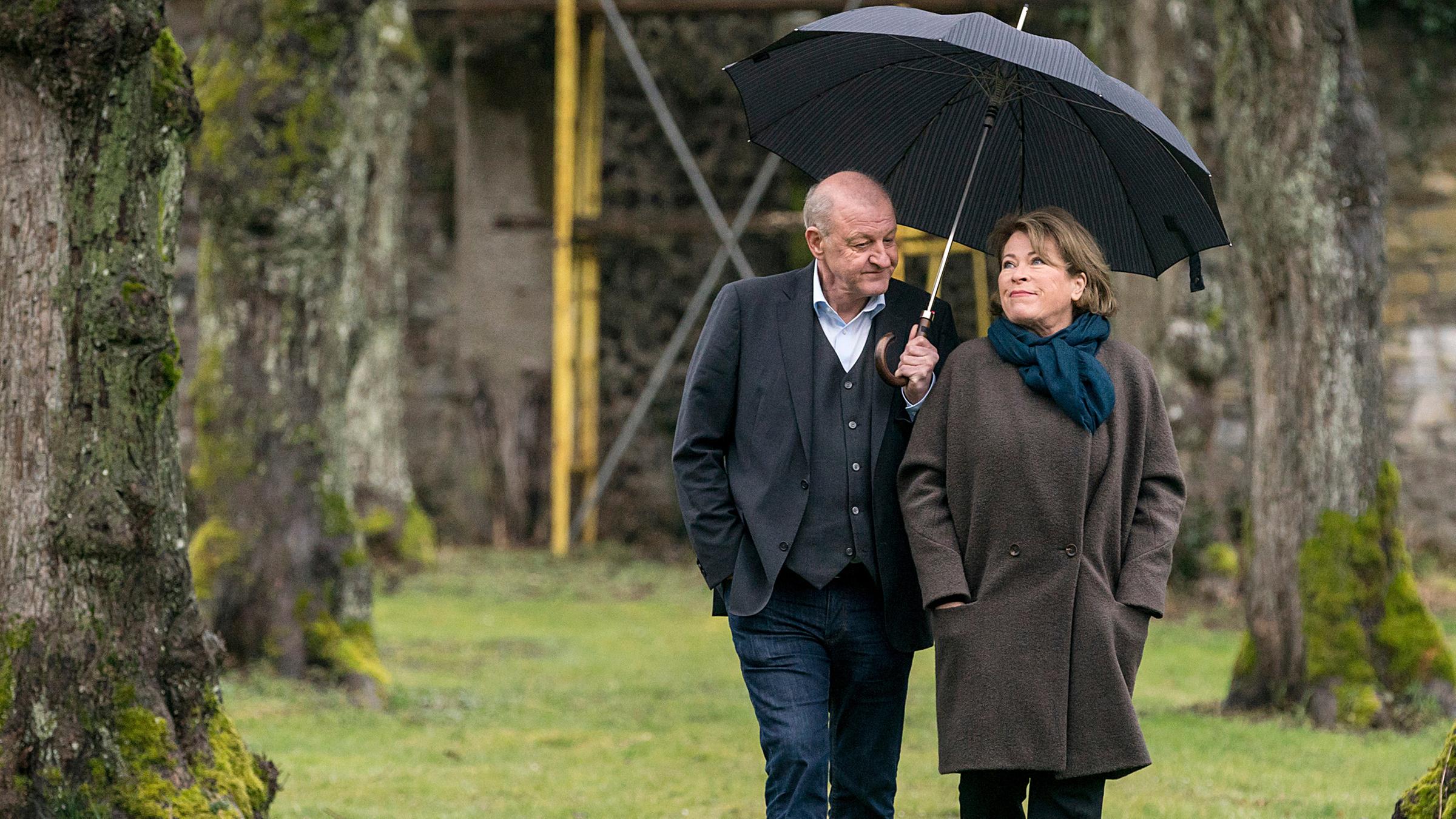 Georg Wilsberg (Leonard Lansink) und Kriminalhauptkommissarin Anna Springer (Rita Russek) gehen, sich einen Regenschirm teilend, im Klosterpark spazieren.