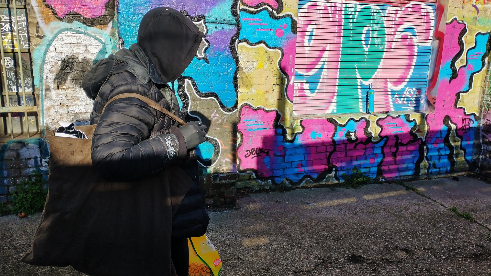 Das Bild zeigt einen Menschen, der schwarz gekleidet ist und eine Kapuze trägt. Man sieht sein Gesicht nicht. In den Händen, die mit schwarzen Handschuhen versteckt sind, trägt er größere Tüten. Er läuft an einer Wand entlang, die voll von bunten Graffitis ist.