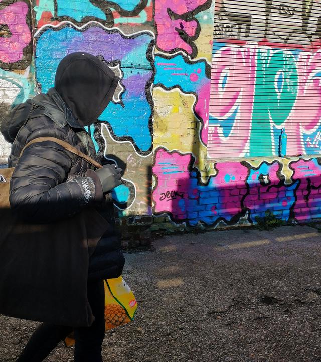 Das Bild zeigt einen Menschen, der schwarz gekleidet ist und eine Kapuze trägt. Man sieht sein Gesicht nicht. In den Händen, die mit schwarzen Handschuhen versteckt sind, trägt er größere Tüten. Er läuft an einer Wand entlang, die voll von bunten Graffitis ist.