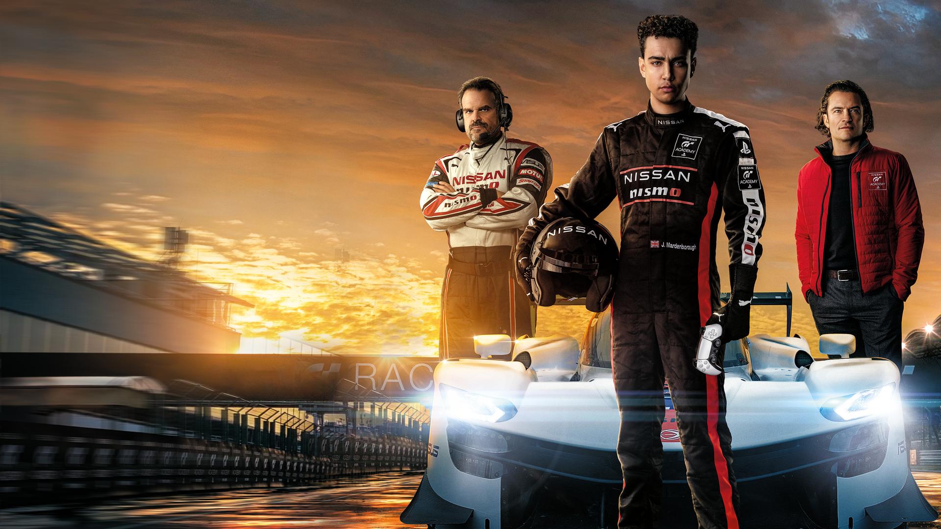 Jann Mardenborough (Archie Madekwe) steht auf einer nächtlichen Rennstrecke im schwarzen Rennanzug und mit dem Helm unter dem Arm vor einem weißen Rennsportwagen. Jack Salter (David Harbour) und Danny Moore (Orlando Bloom) posieren neben dem Wagen. 