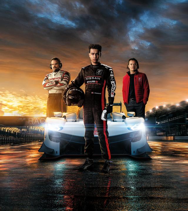 Jann Mardenborough (Archie Madekwe) steht auf einer nächtlichen Rennstrecke im schwarzen Rennanzug und mit dem Helm unter dem Arm vor einem weißen Rennsportwagen. Jack Salter (David Harbour) und Danny Moore (Orlando Bloom) posieren neben dem Wagen. 