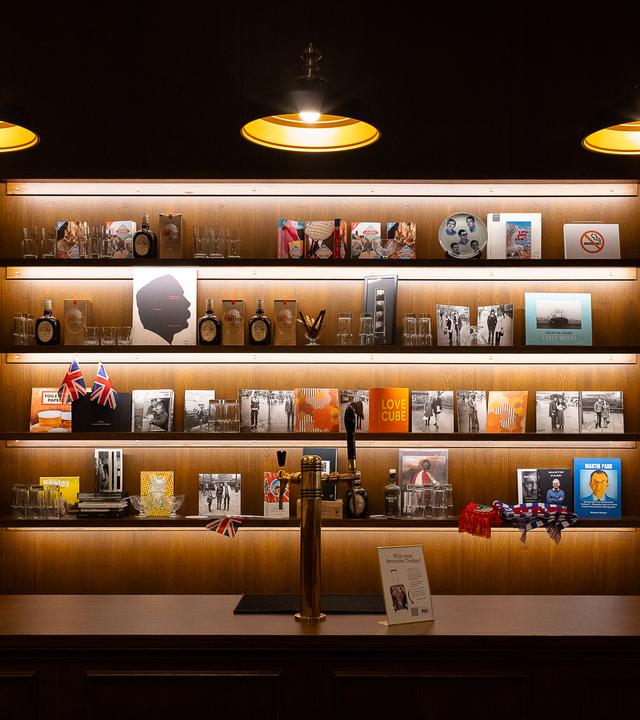 Eine Theke mit Zapfhahn und dahinter ist ein Bücherregal, in dem verschiedene Bilder, Bücher, Parfums und auch Flaggen stehen. Von oben hängen drei Lampen runter ins Bild.