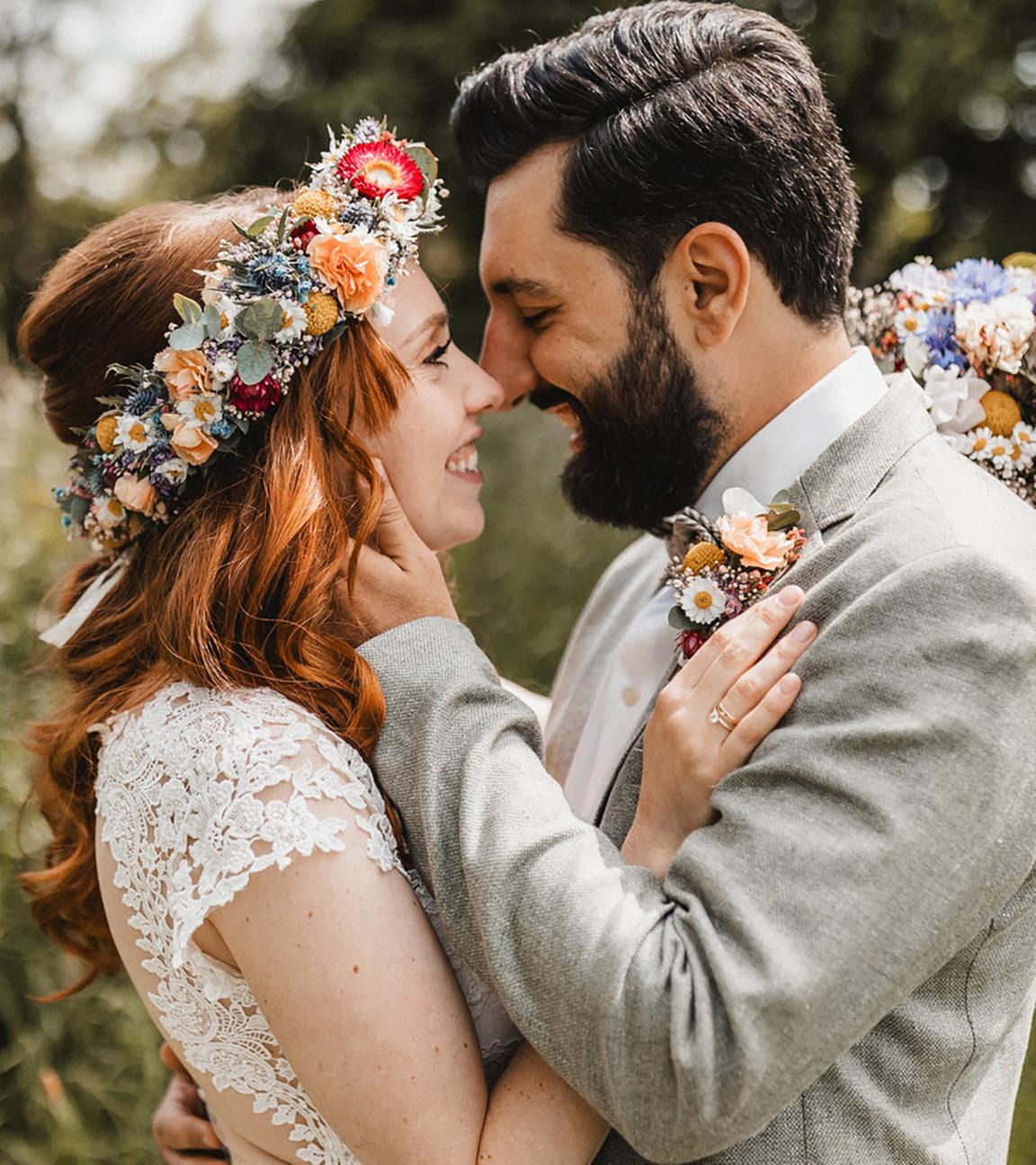 Im Grünen: Ein Brautpaar schaut sich tief in die Augen und lacht sich an und scheint sich gleich küssen zu wollen. Janina Rehm trägt lange, rote, offene Haare, die von einem Blumenkranz geschmückt sind. Ihre Hand hat sie auf die Brust ihres Mannes gelegt, der einen grauen Anzug trägt. Er hat einen dichten, schwarzen Vollbart und hält mit seiner linken Hand zärtlich den Nacken seiner Frau. Die Braut hält einen bunten Blumenstrauß in der Hand, mit der sie ihren Mann umarmt. Merter trägt ein kleines Blumengesteck am Revers seines Anzugs.