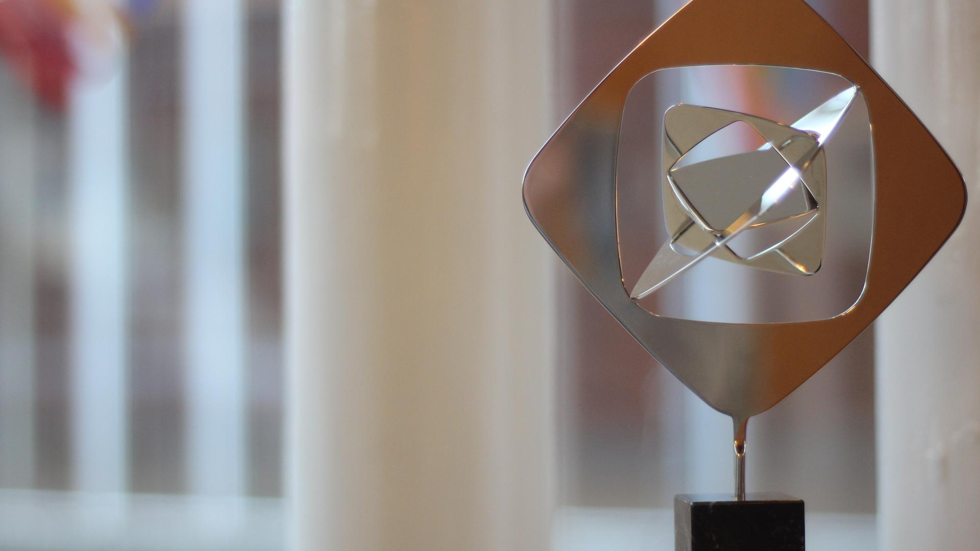 Das Bild zeigt eine moderne, abstrakte Skulptur aus poliertem Metall, die auf einem schwarzen Sockel steht, vor einem unscharfen Hintergrund eines Fensters mit weißen Vertikalstreben. 
