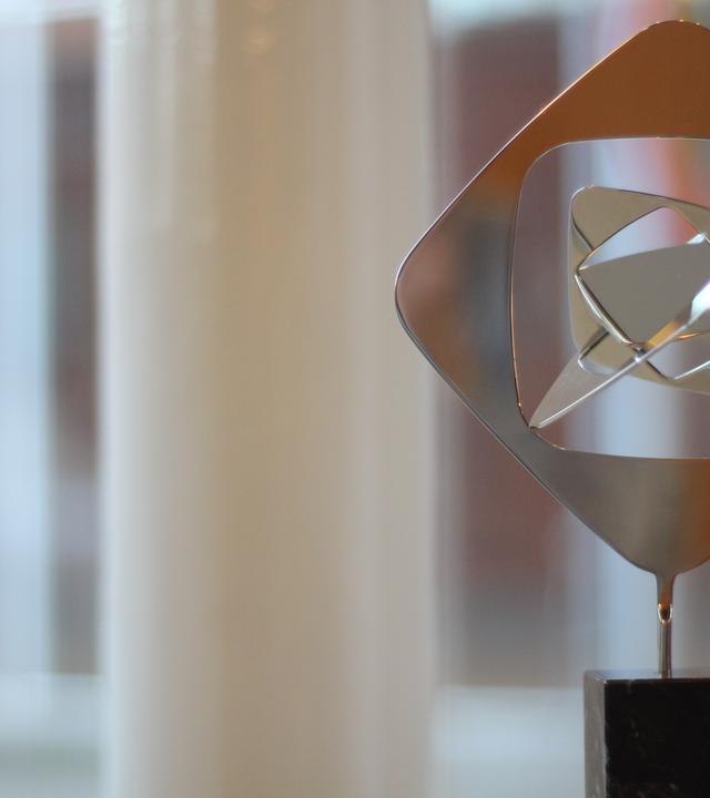 Das Bild zeigt eine moderne, abstrakte Skulptur aus poliertem Metall, die auf einem schwarzen Sockel steht, vor einem unscharfen Hintergrund eines Fensters mit weißen Vertikalstreben. 