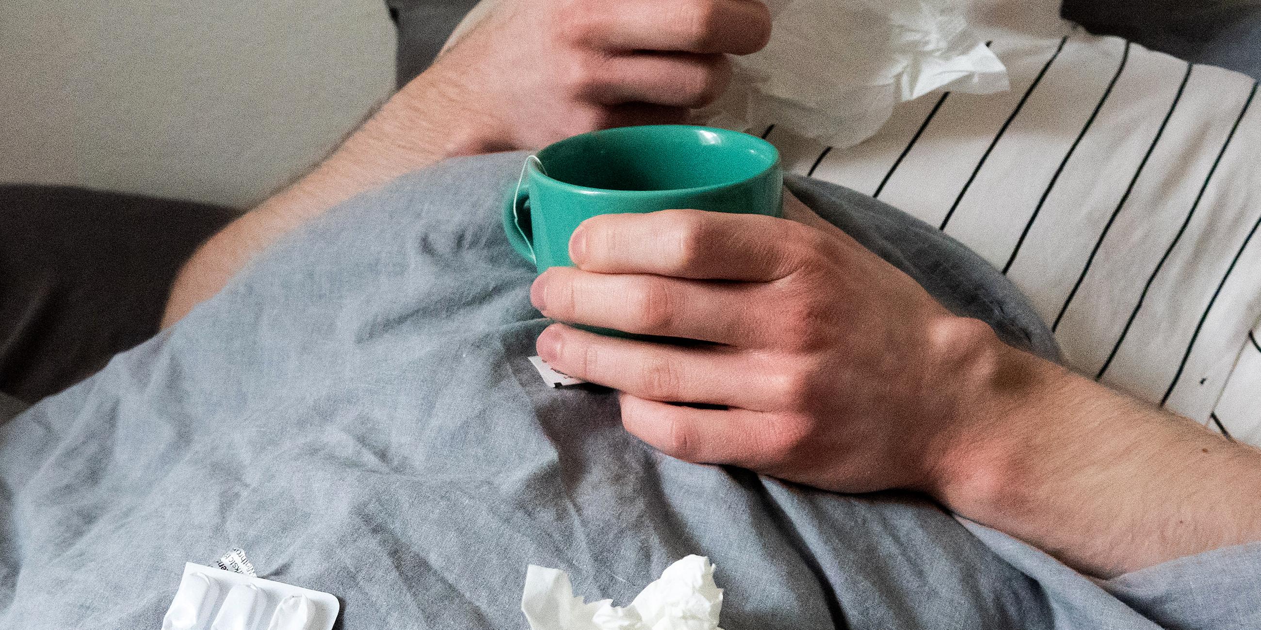Symbolbild: Ein Mann liegt mit Taschentüchern, Teetasse, Nasenspray und Tabletten im Bett (gestellte Szene).